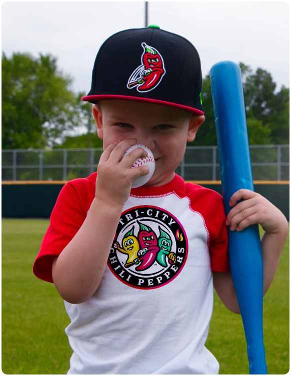 Young baseball fan wearing a Tri-City Chili Peppers jersey and hat, holding a baseball and blue bat at the stadium.