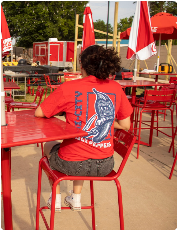 Model sitting at a stadium table wearing a red Tri-City Chili Peppers t-shirt with a blue and white mascot logo on the back.