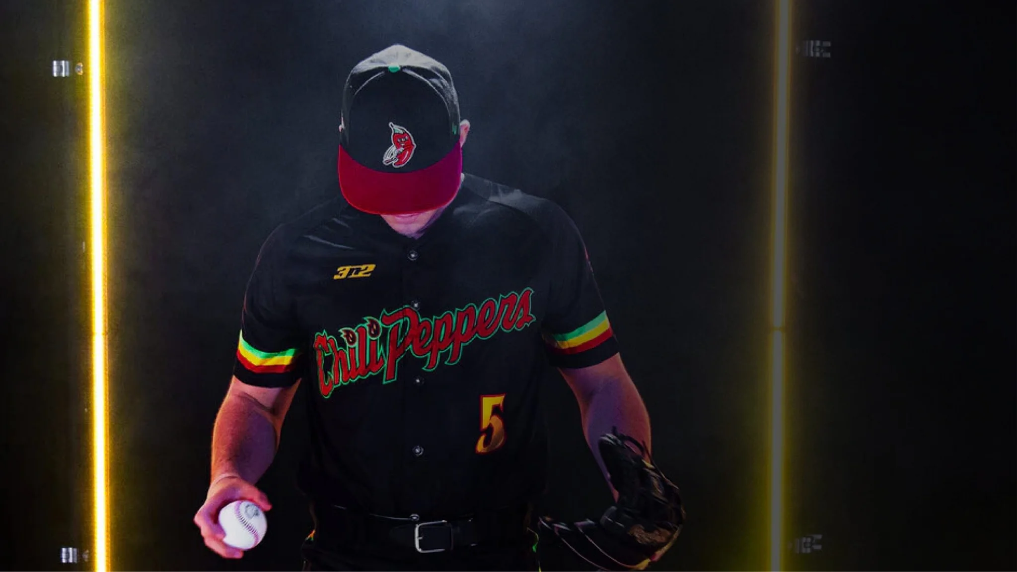Tri-City Chili Peppers pitcher in a black jersey and red pepper cap holding a baseball, framed by glowing vertical lights.