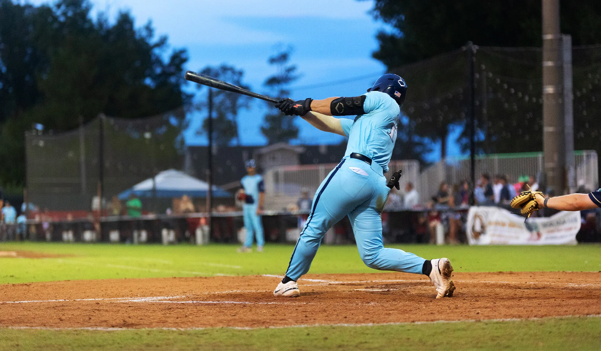 Baseball player in a powder blue uniform swinging a bat during a night game with trees and fans in the background.